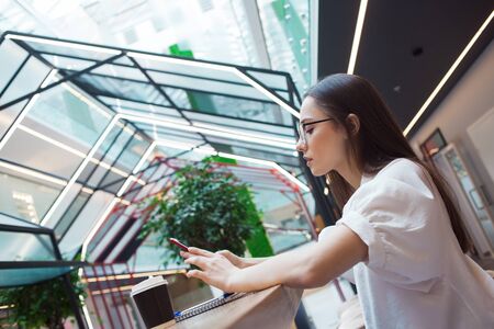young beautiful woman in glasses and a white blouse is sitting in a public space and looking at the screen of a smartphone. The girl in the coworking works online, social media Manager and freelancerの写真素材