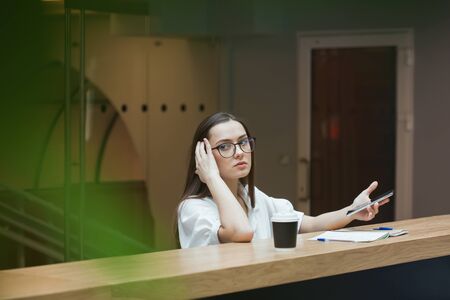 young beautiful woman in glasses and a white blouse is sitting in a public space and looking at the screen of a smartphone. The girl in the coworking works online, social media Manager and freelancerの写真素材