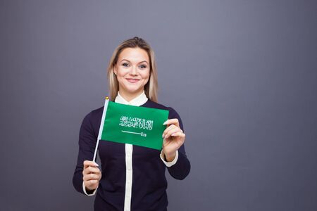 Immigration and the study of foreign languages, concept. A young smiling woman with a Saudi Arabia flag in her hand. Girl waving a Saudi Arabia flag on a gray backgroundの写真素材