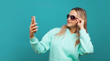 Stylish trendy hipster girl in the blue hoodie with the hood uses a smartphone. Happy young blonde enjoys social networking and a good appの写真素材
