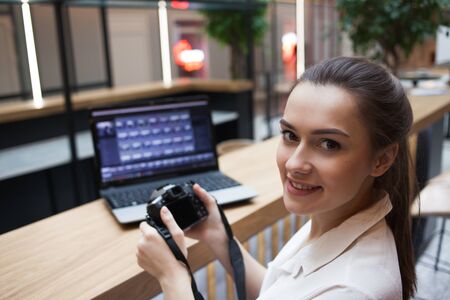 Cute young girl working at a laptop and holding a camera in her hands. Blogger and photographer,...