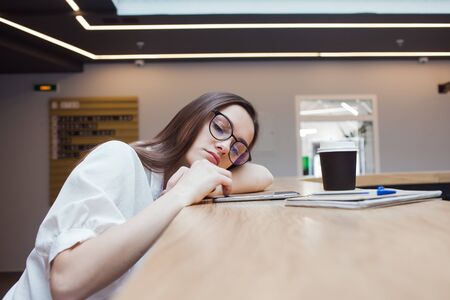 tired young female student sits at a high table top and is sad with a phone in her hands. Young...