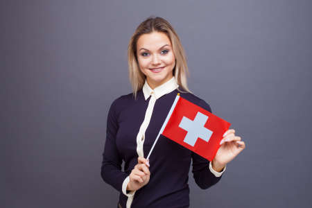 Immigration and the study of foreign languages, concept. A young smiling woman with a Switzerland flag in her hand. Girl waving a Swiss flag on a gray backgroundの写真素材