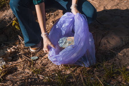 Cleaning up garbage, volunteer work. Caring for nature. People left a lot of plastic garbage in the forest. A young woman collects plastic waste in a biodegradable bag. package, close-upの写真素材