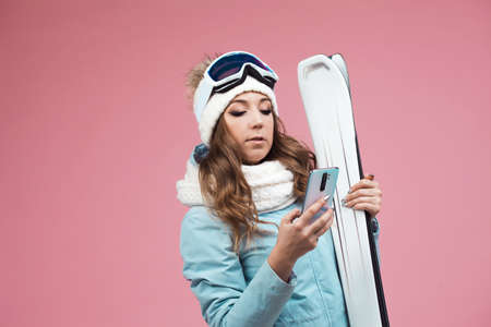 young woman with skis uses a smartphone to plan her ski runs. Winter holidays at a ski resort. Girl with a hat and skis on a pink backgroundの写真素材