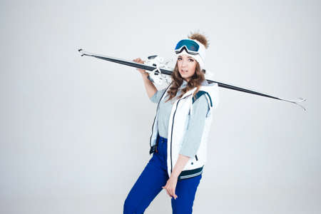 Smiling girl skier in a hat and mask for skiing. A young woman in clothes for skiing and outdoor activities. A woman holds skis and poses on a white background in the Studio.の写真素材