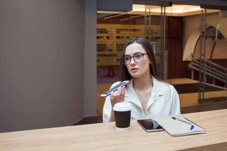 The girl in the coworking works online, a young business woman with glasses uses voice input or sends a voice message in a messenger. Modern communication in a smartphoneの写真素材