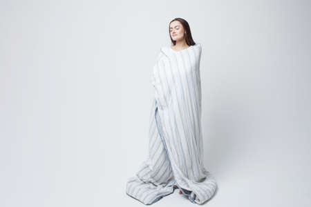 Healthy sleep and drowsiness, beautiful young woman standing wrapped in a blanket, light gray...