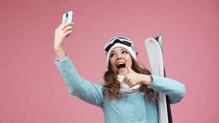 fun skier takes a selfie on her smartphone. A young woman with ski equipment in a jacket and hat, a...