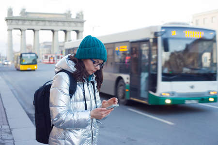 woman in a down jacket and hat stands at a public transport stop and uses a smartphone. Wait for the...