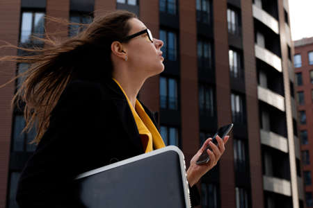 young business woman with a laptop and smartphone goes to work. Modern business lady in casual coat and glasses on the background of the business centerの写真素材