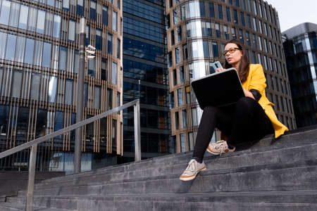 young woman in a bright yellow jacket uses a laptop and smartphone. freelancer, remote worker, or...