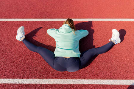 Training on the street, a young female athlete makes a workout on the street platform. Do stretching while sitting on the ground, outdoor surface for sports fields and treadmillsの写真素材