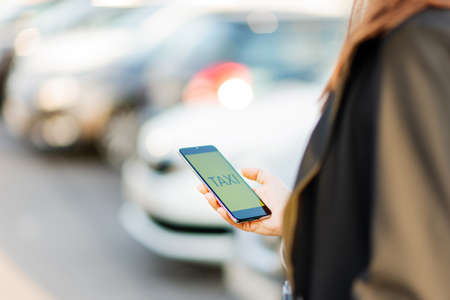 Call a taxi using the mobile app on your smartphone. A young woman is waiting for a taxi standing by the road, holding a phone with a mobile serviceの写真素材