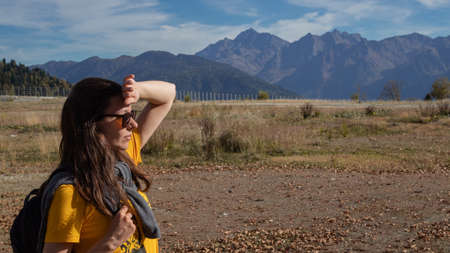 young female tourist with a backpack stands in a mountain valley against the background of peaks,...