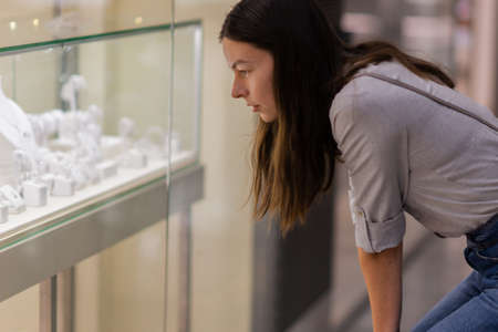 A young woman in a store admires a display of jewelry with diamonds and precious stones. Jewelry storeの写真素材