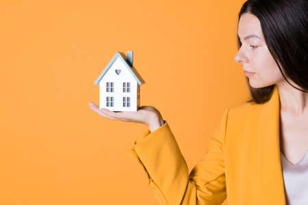 Buy a house, take out a Bank loan or save up your money. Real estate investment, concept. A young woman in yellow on a yellow background holds a house in her hands.の写真素材