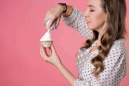 young charming female pastry chef decorates cupcakes with whipped cream. A cook in a polka-dot...