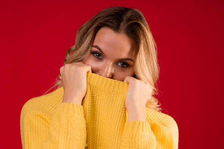 young sweet and smiling blonde woman in a yellow knitted sweater on a red background, wrapped in a collarの写真素材