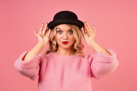 A young, attractive woman in a warm pink sweater and a black bowler hat on a pink background, blonde with curly hair and red lipstickの写真素材
