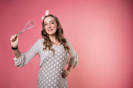 young charming pastry chef woman with a whisk in her hands. Do magic in the kitchen, wave a whisk like a magic wand. A cook in a polka-dot apron. pastry chef in an apron on a pink background.の写真素材