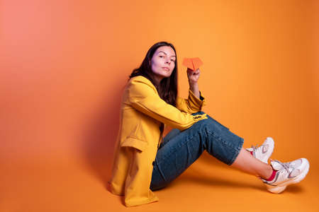 Launch a paper airplane, a dream travel concept. Young brunette woman in yellow sitting on a yellow background, studio shot.の写真素材