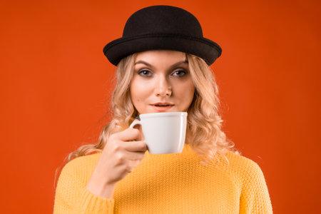 Espresso or Americano, a young stylish blonde in a bowler hat, holding a cup in her hands. Enjoy coffee, studio portrait on orange backgroundの写真素材