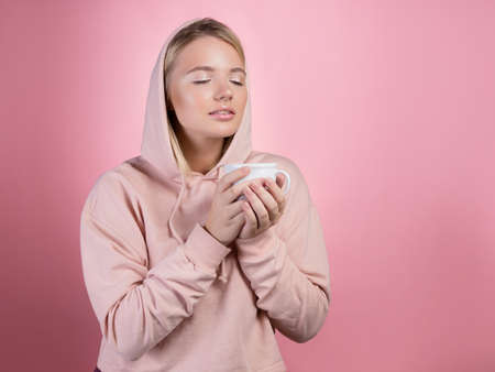 Enjoy a warming drink, a young sweet woman in a pink hoodie holds a mug