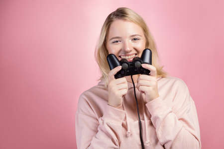 Charming blonde gamer. A young smiling woman in a pink hoodie holds a joystick from a game console in her hands, winning the round. Portrait on a pink backgroundの写真素材