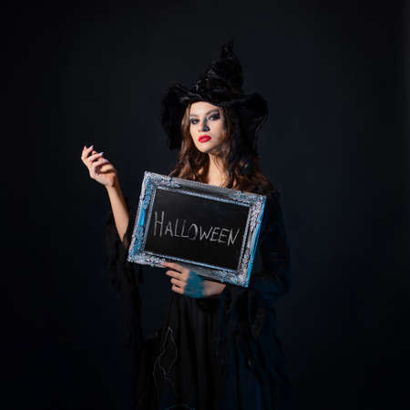 A young beautiful witch in a pointed hat holds a sign with the inscription Halloween, an invitation to a costume party.の写真素材