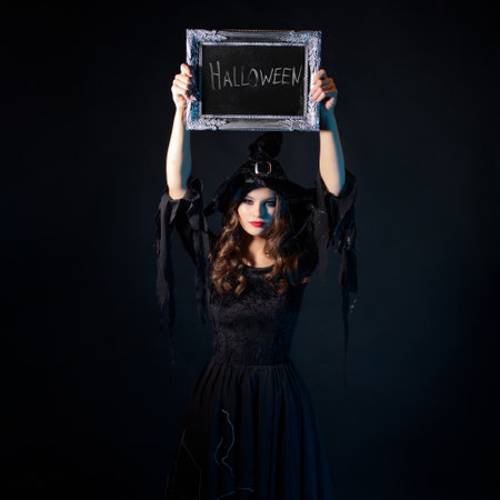A young beautiful witch in a pointed hat holds a sign with the inscription Halloween, an invitation to a costume party.の写真素材