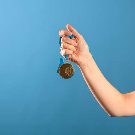 Win ,be a champion. Striving for achievements, concept, a female hand squeezes the winners gold medal, blue backgroundの写真素材