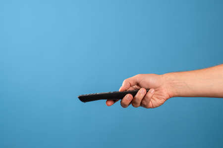 Watch new TV series and movies at home on streaming services, the TV remote control is in a mans hand, switch channels. Photo on a blue backgroundの写真素材
