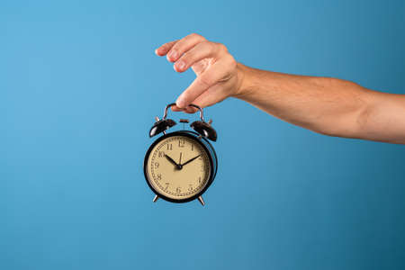 Wake up and get up, its time to act, concept. Clock and alarm clock, attention its time to do. A mans hand holds a vintage alarm clock, a photo on a blue backgroundの写真素材
