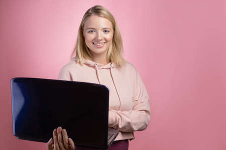young woman with a laptop, a student or a freelancer. A charming blonde in a pink hoodie on a pink background holds a laptop in her hands.の写真素材