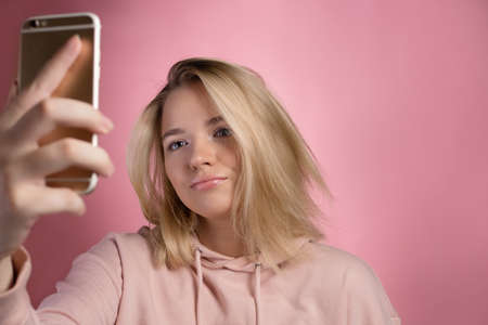 attractive young woman in a pink hoodie uses a smartphone for video communication or photos. Cute blonde, photo on pink.の写真素材