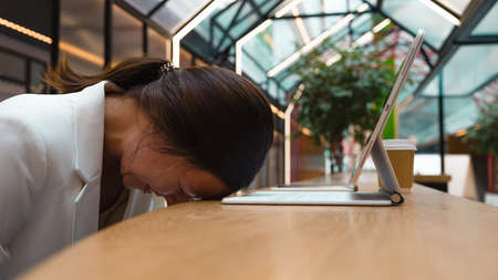 Professional burnout in a freelancer, chronic fatigue and panic attacks when working remotely. A young woman lies with her head on the table in front of a laptop and laughs nervouslyの写真素材