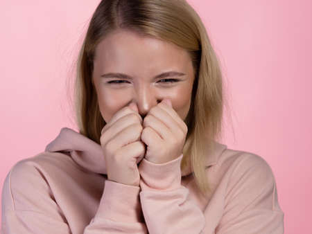 Cute young blonde in a pink hoodie squints, confused face. portrait on a pink background, close-upの写真素材