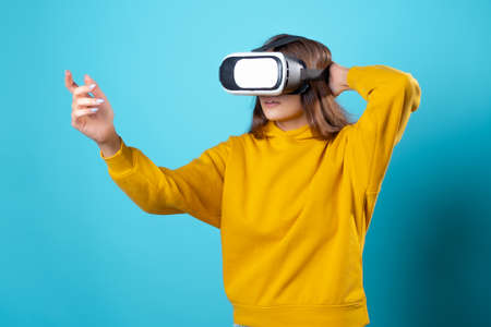 VR gamer, a young woman in a youth outfit uses a virtual reality helmet. Brunette in a yellow hoodie on a blue background with a virtual reality helmet on her head, full immersionの写真素材