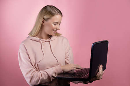young woman, a student or a freelancer, uses a laptop for work or study. Blonde in a pink hoodie on a pink backgroundの写真素材