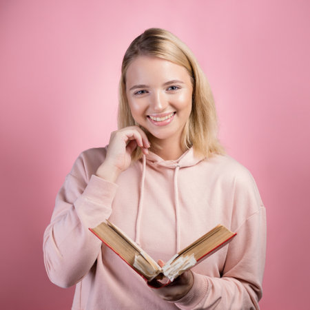 A young woman in a pink hoodie is studying a textbook with a lot of bookmarks, an open book in her hands, pink backgroundの写真素材