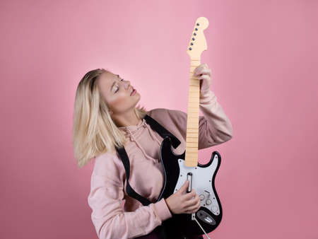 A young charming blonde is laughing and playing a toy electric guitar, fun and hobby. A cheerful young woman in a pink hoodie on a pink background. Enjoy music, soloの写真素材