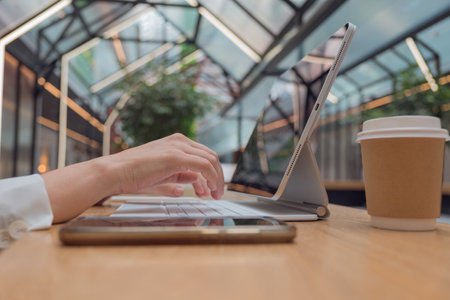 Remote work or freelance, modern opportunities for a mobile lifestyle. Womens hands are typing on keyboard of tablet computer, close-up side view, public space hall on background, low depth of field.の写真素材