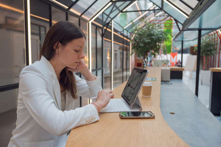 A real estate specialist works remotely. A young woman in a white jacket sits in front of a laptop screen and looks at an engineering floor plan, development or study of the layout.の写真素材