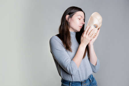 A young woman hides her face behind a mask, the concept of psychological problems, a crisis of self-identification or strong shyness. Photos in light gray tones, gray background.の写真素材