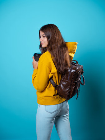 A young student in a yellow hoodie with a cardboard cup of coffee to take away. A girl with a backpack, Portrait on a blue backgroundの写真素材