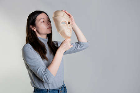A young woman hides her face behind a mask, the concept of psychological problems, a crisis of self-identification or strong shyness. Photos in light gray tones, gray background.の写真素材