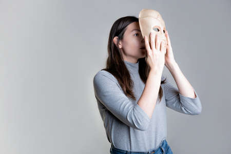 Complexes due to appearance or self-identification problems, concept. A young woman holds a mask in her hands hiding her face. Photos in light gray tones, gray backgroundの写真素材