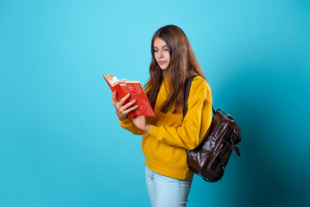 A student with a book in her hands, reading a textbook and preparing for a new lesson. A young brunette in a yellow hoodie and with a backpack, leafing through a book.の写真素材