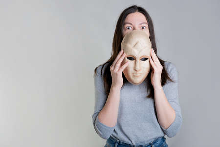 A young woman holds a mask in her hands, hiding her face half, a surprised look over the mask. Photos in light gray tones, gray backgroundの写真素材